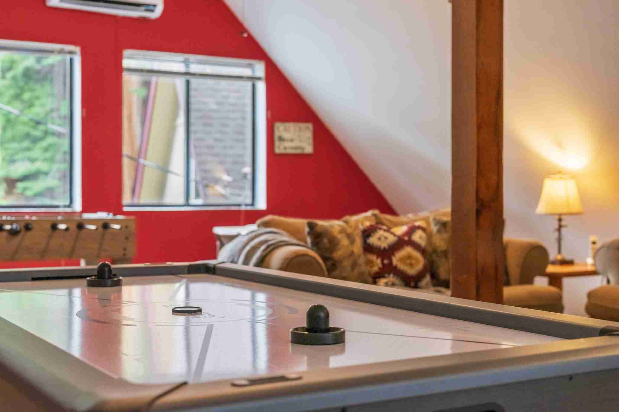 Cozy room with air hockey table, foosball, seating area, red wall, and natural light from windows.