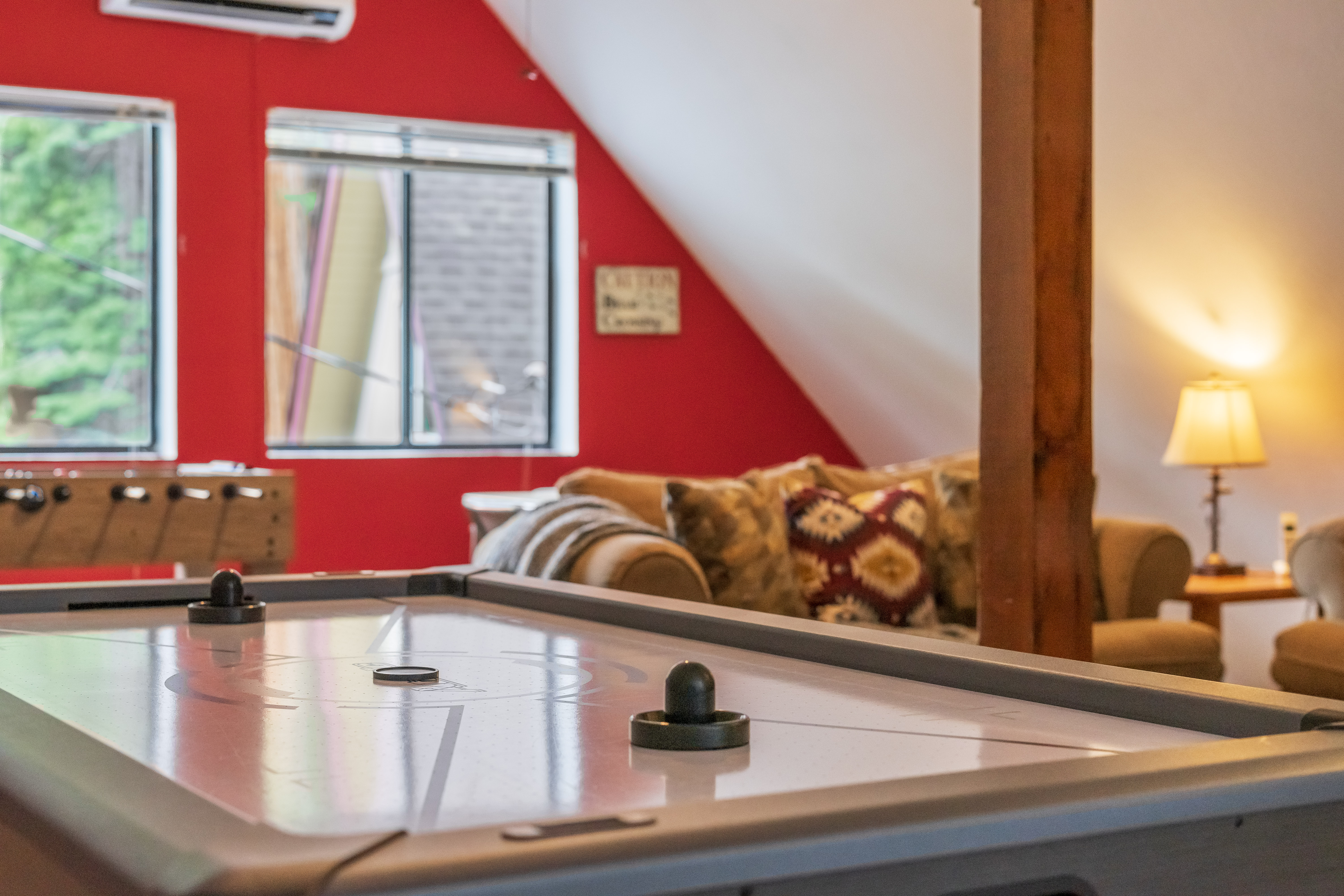 Cozy room with air hockey table, foosball, seating area, red wall, and natural light from windows.