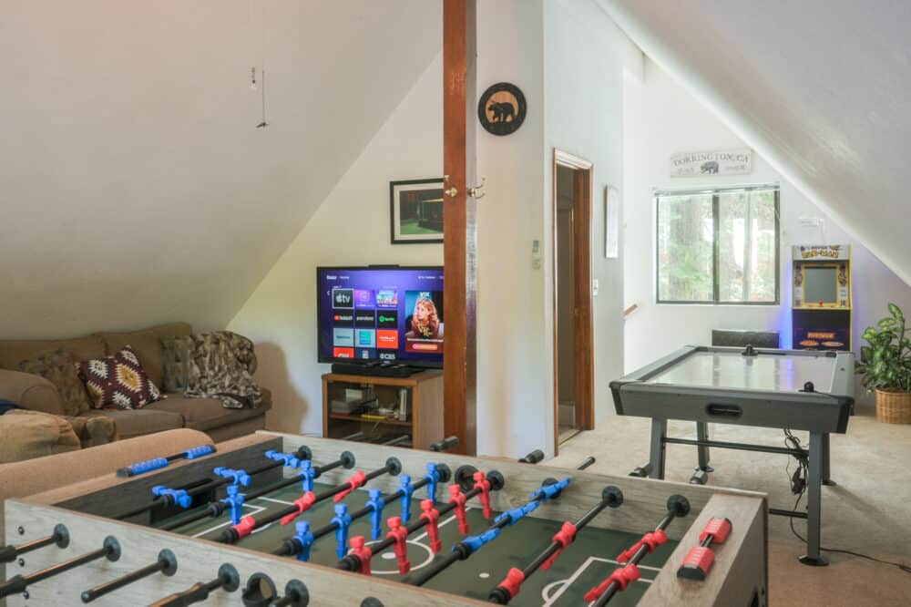Dorrington Vacation Rental Cabins - A bright game room with a foosball table, a tv showing a streaming service menu, and an air hockey table under a vaulted ceiling.