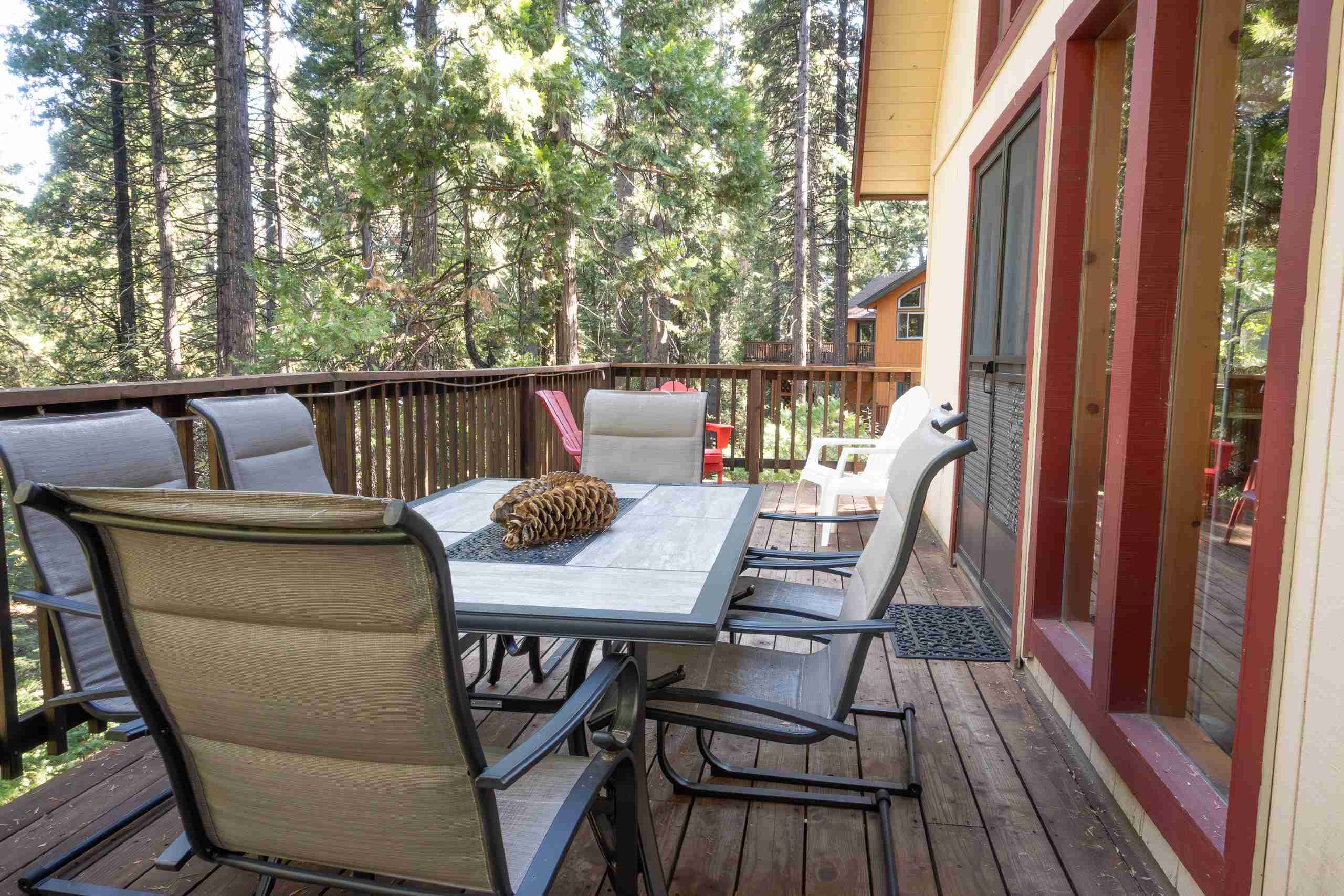 Wooden deck with dining table, cushioned chairs, pine cone decor, forest view, and large windows.