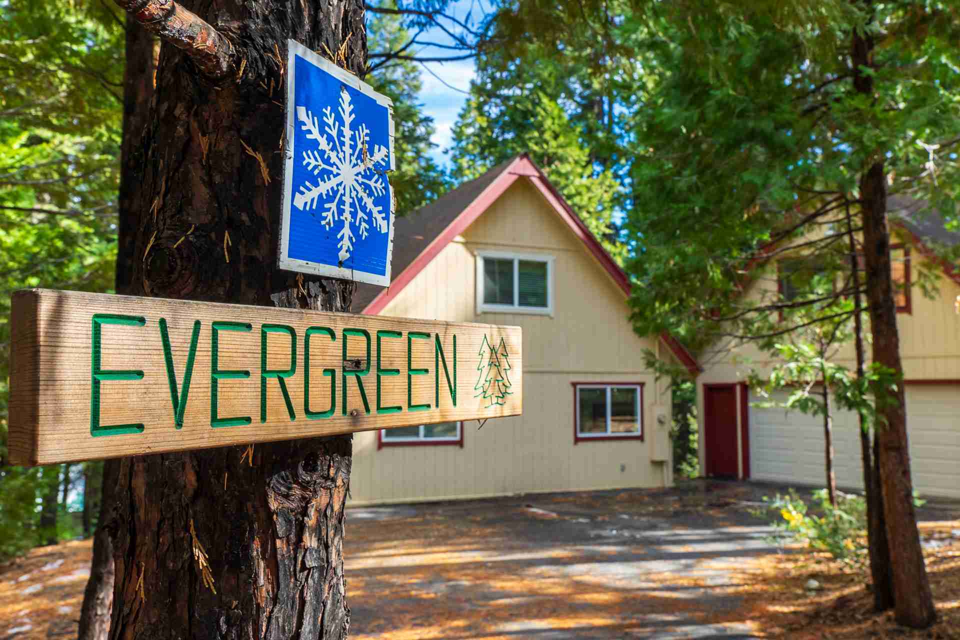 Wooden "Evergreen" sign on tree with snowflake, near house with maroon roof and pine trees.