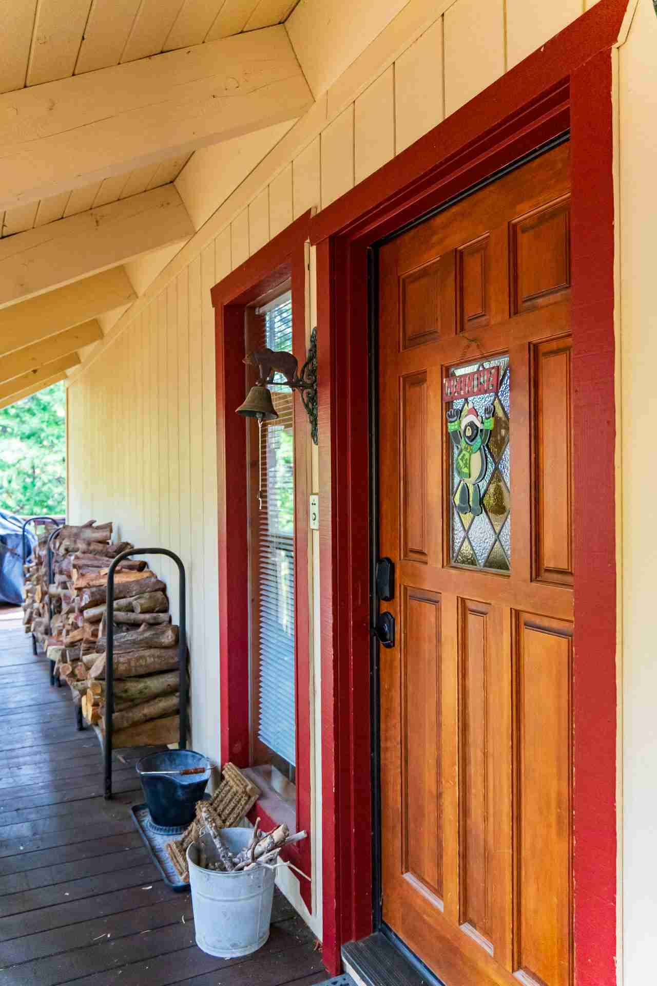 Wooden porch with red-framed door, bell, wood stack, blinds, and galvanized buckets.
