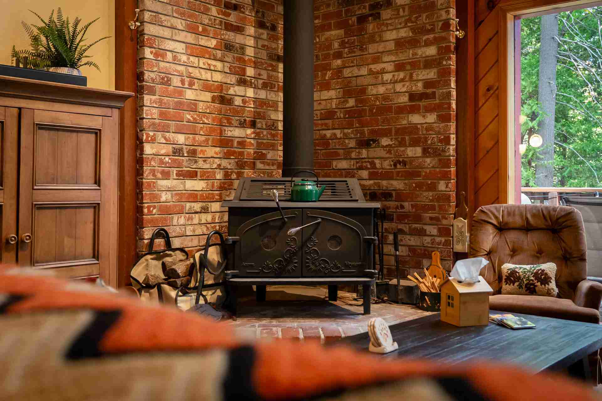Cozy living room with brick fireplace, wood stove, green kettle, chair, and window view of trees.