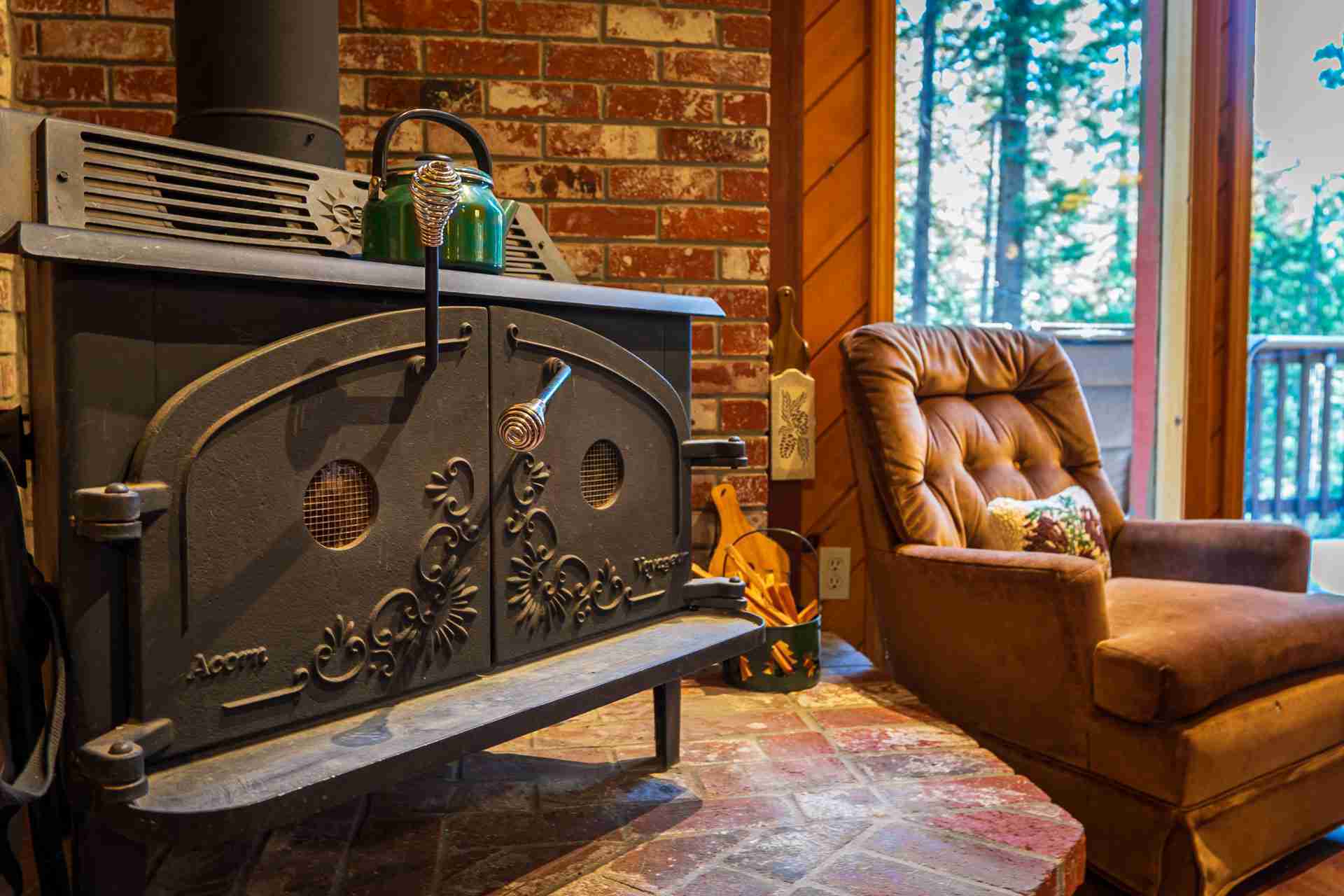 Wood stove with a green kettle, armchair nearby, brick wall, large window view of trees.