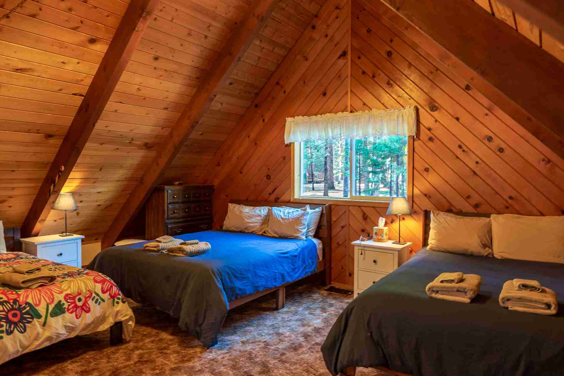 Cozy attic bedroom with wooden walls, sloping ceiling, two beds, lamps, dresser, and window.