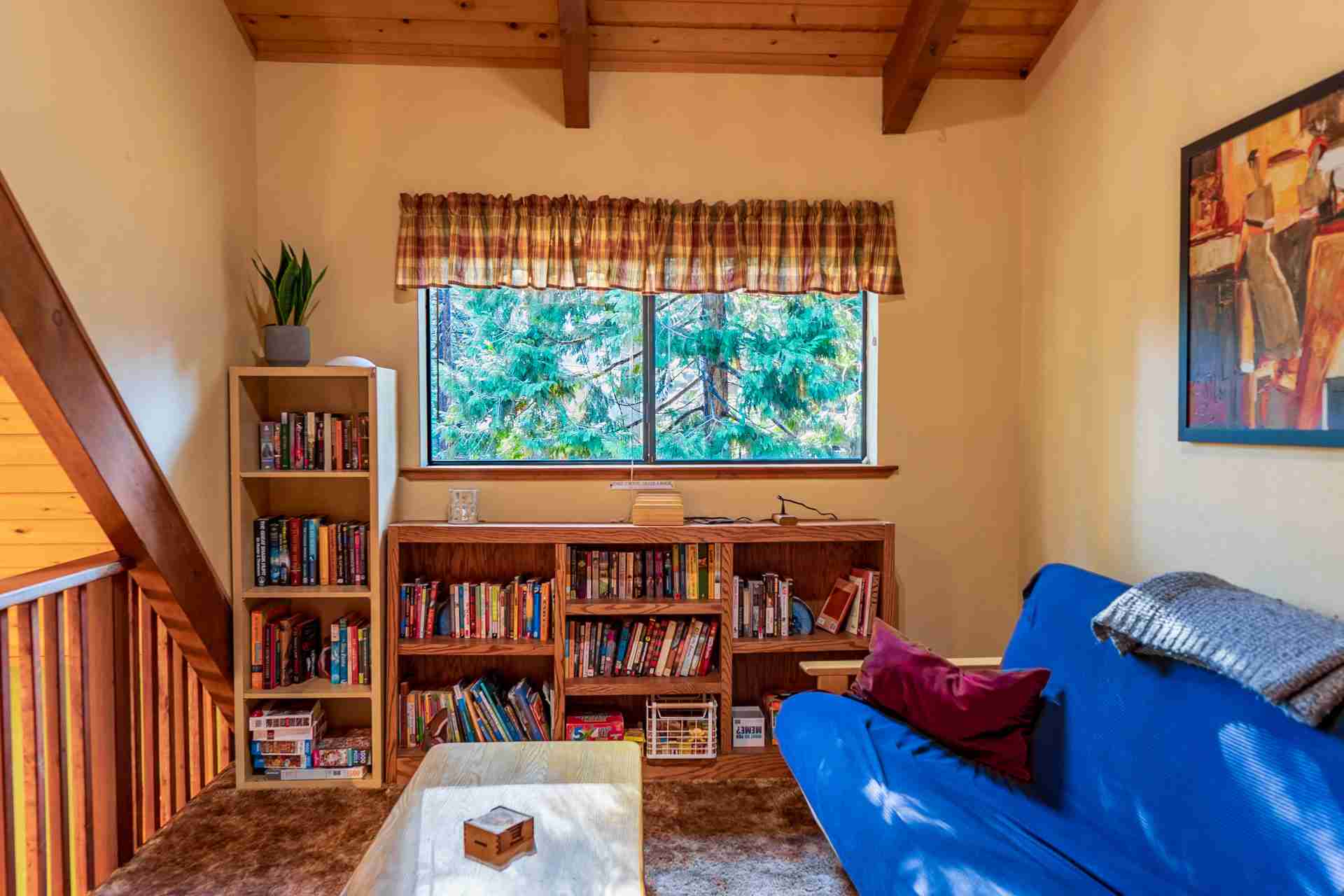Cozy room with wooden ceiling, blue sofa, bookshelves, window showing greenery, and painting.