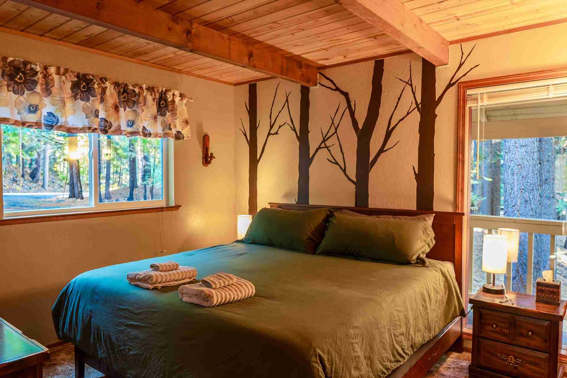 Cozy bedroom with wooden beams, green bed, tree motif wall, floral curtains, and window view.