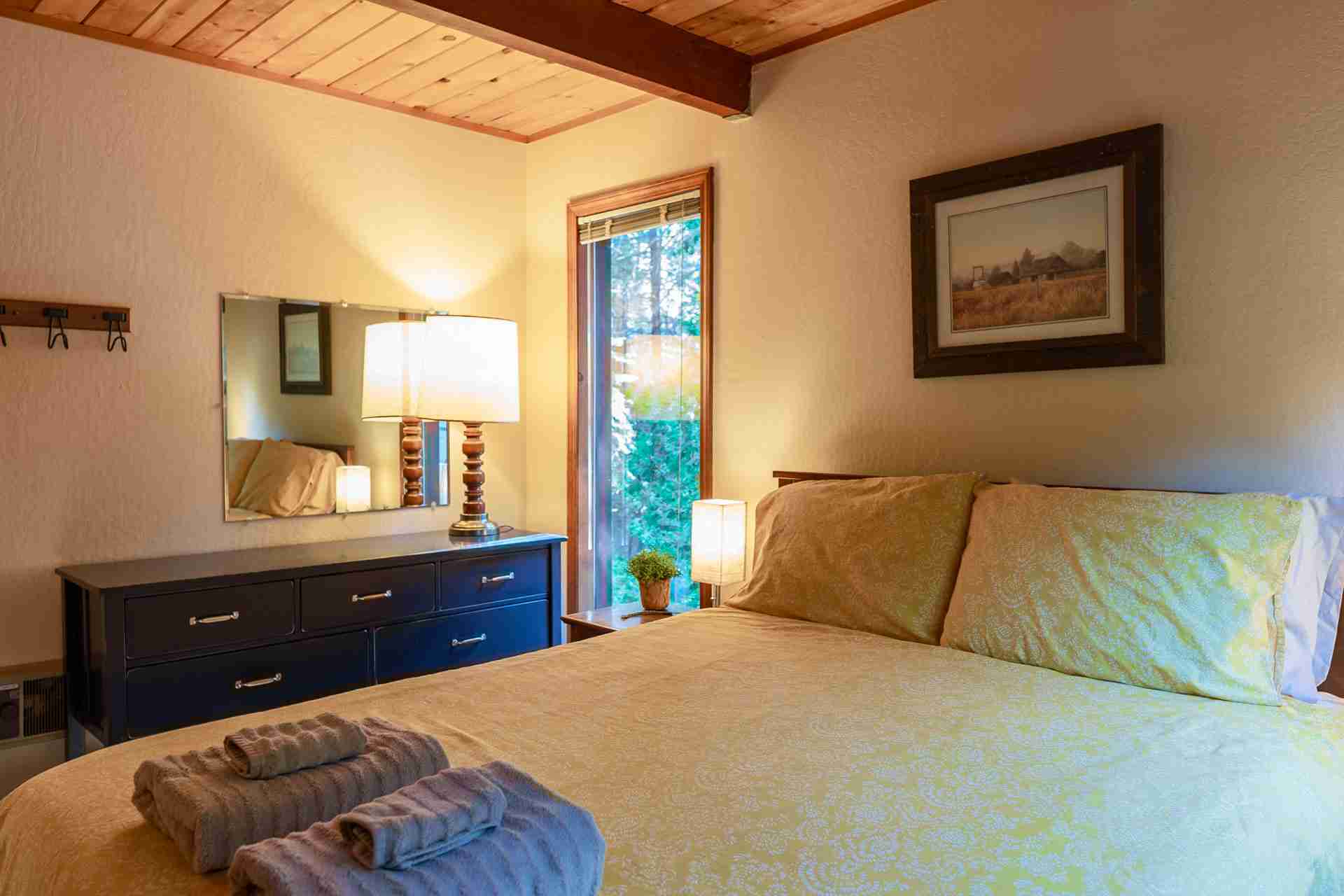 Bedroom with yellow bedding, towels, wooden dresser, lamp, mirror, picture, window, ceiling beams.
