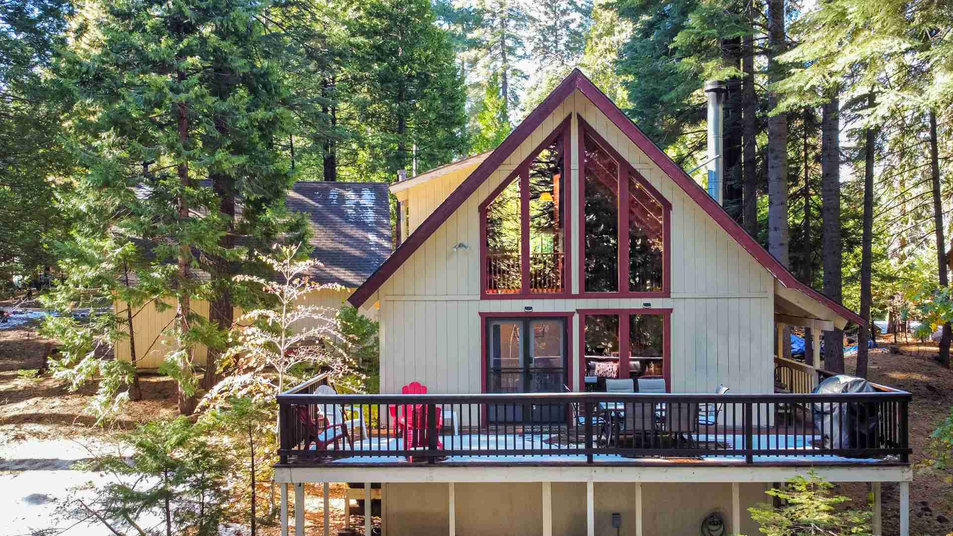 Two-story A-frame cabin with large windows, wooden deck, in forested area with tall trees.