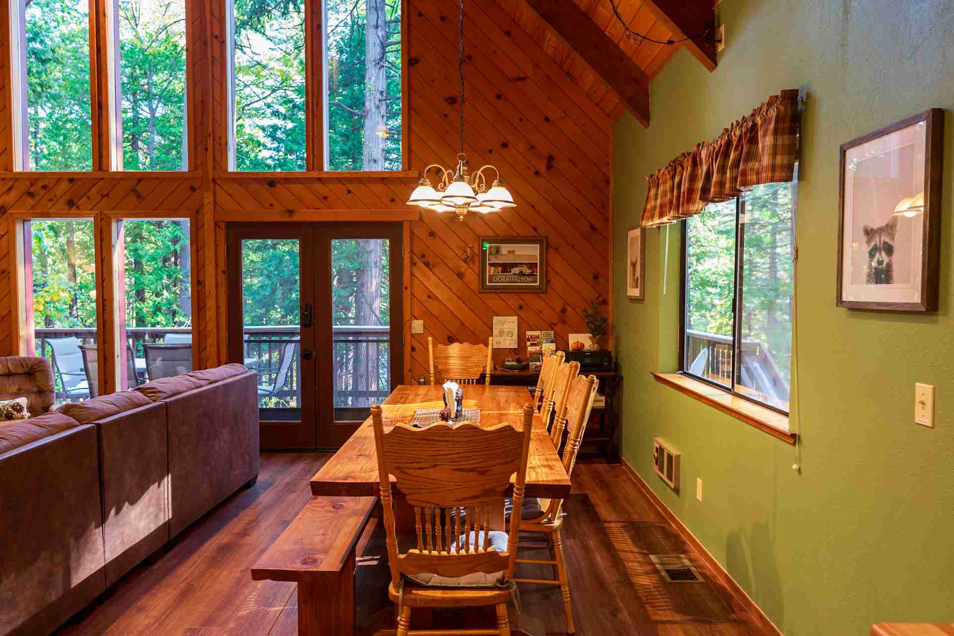 Cozy cabin dining area with wooden table, chairs, sofa, large windows, and outdoor view.